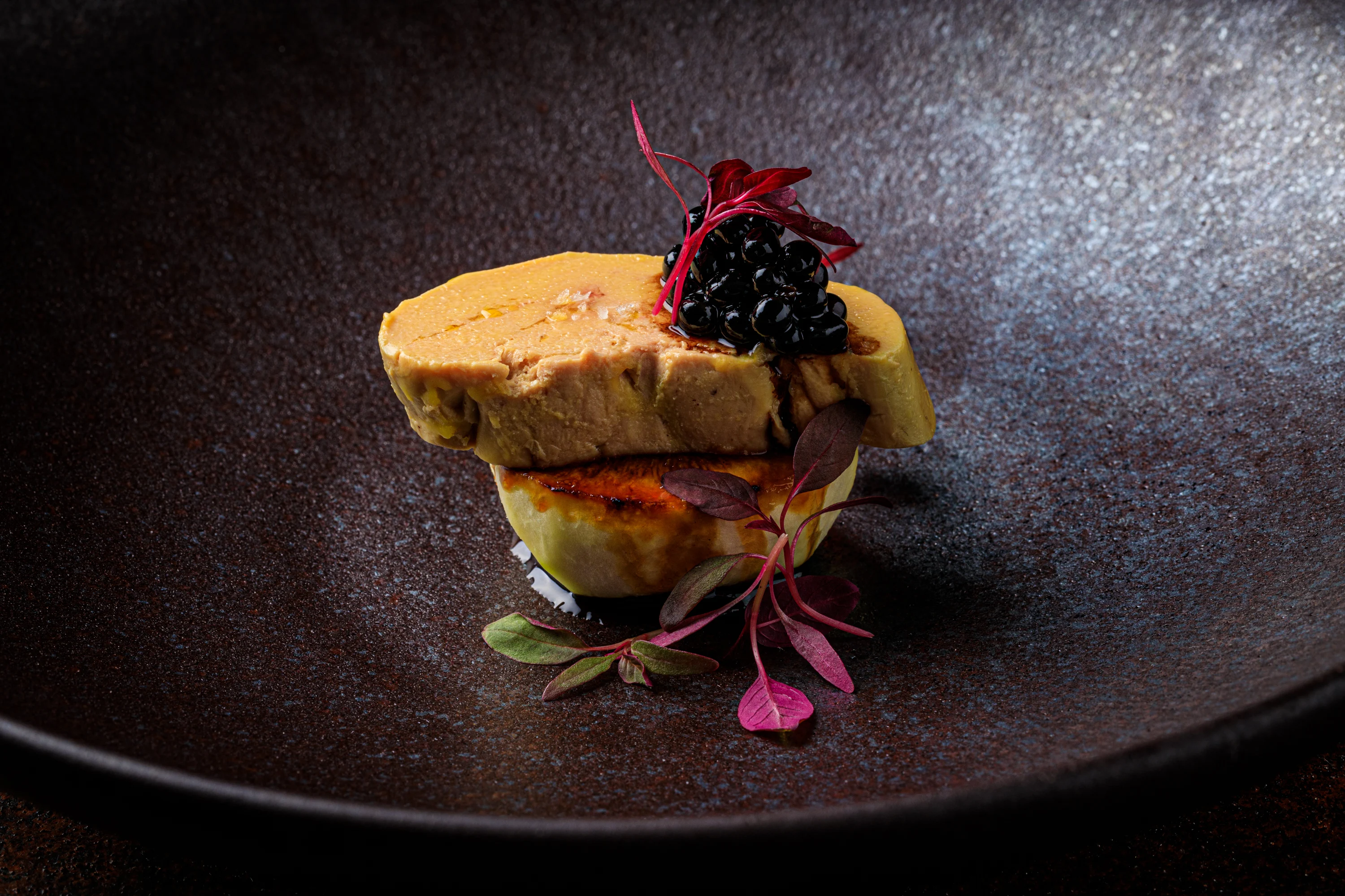 A layered foie gras course with dark berry garnish on a textured plate.