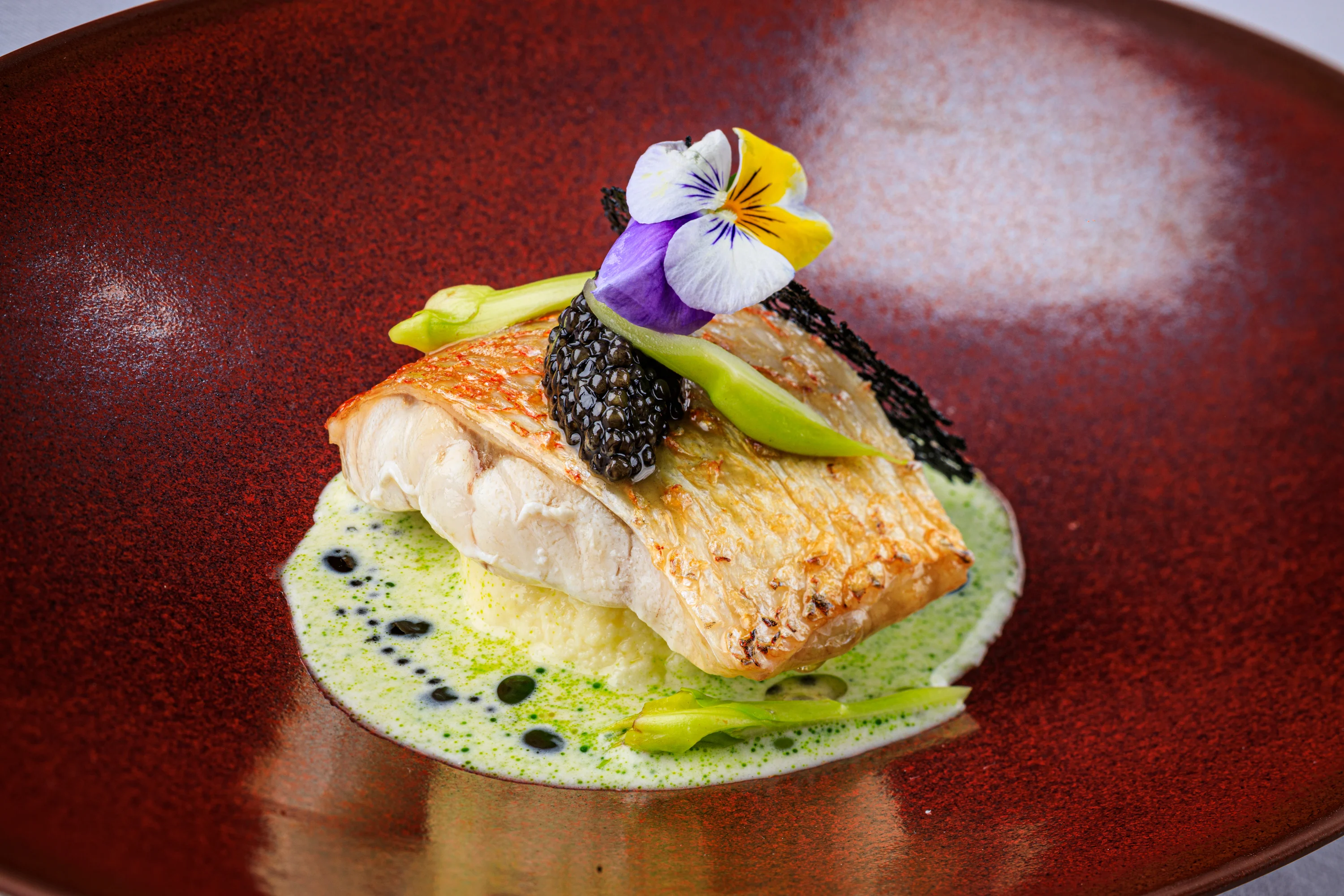 A fish course with caviar, flowers and pale green sauce on a red plate.