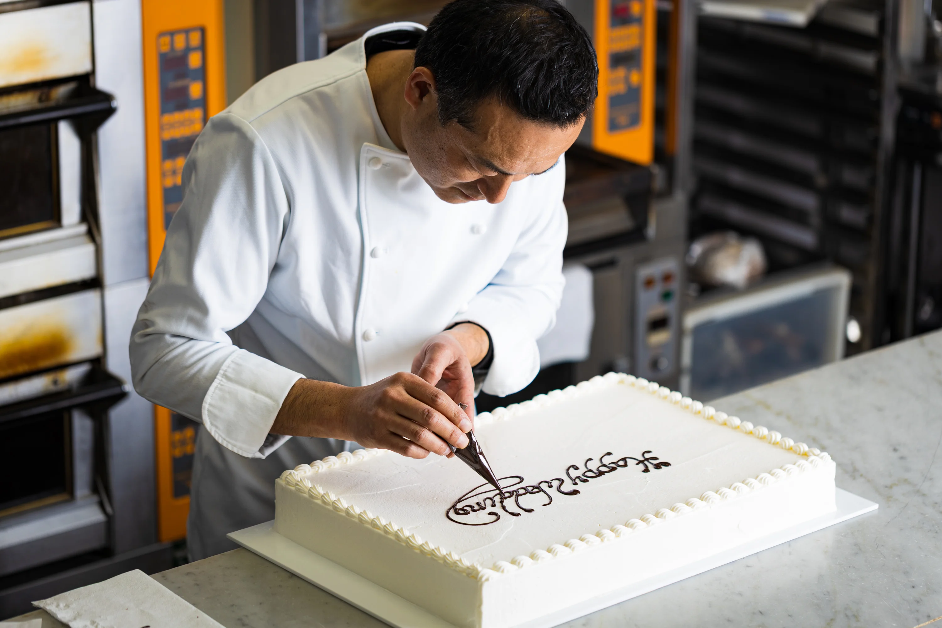 A chef piping chocolate lettering onto a large celebration cake.