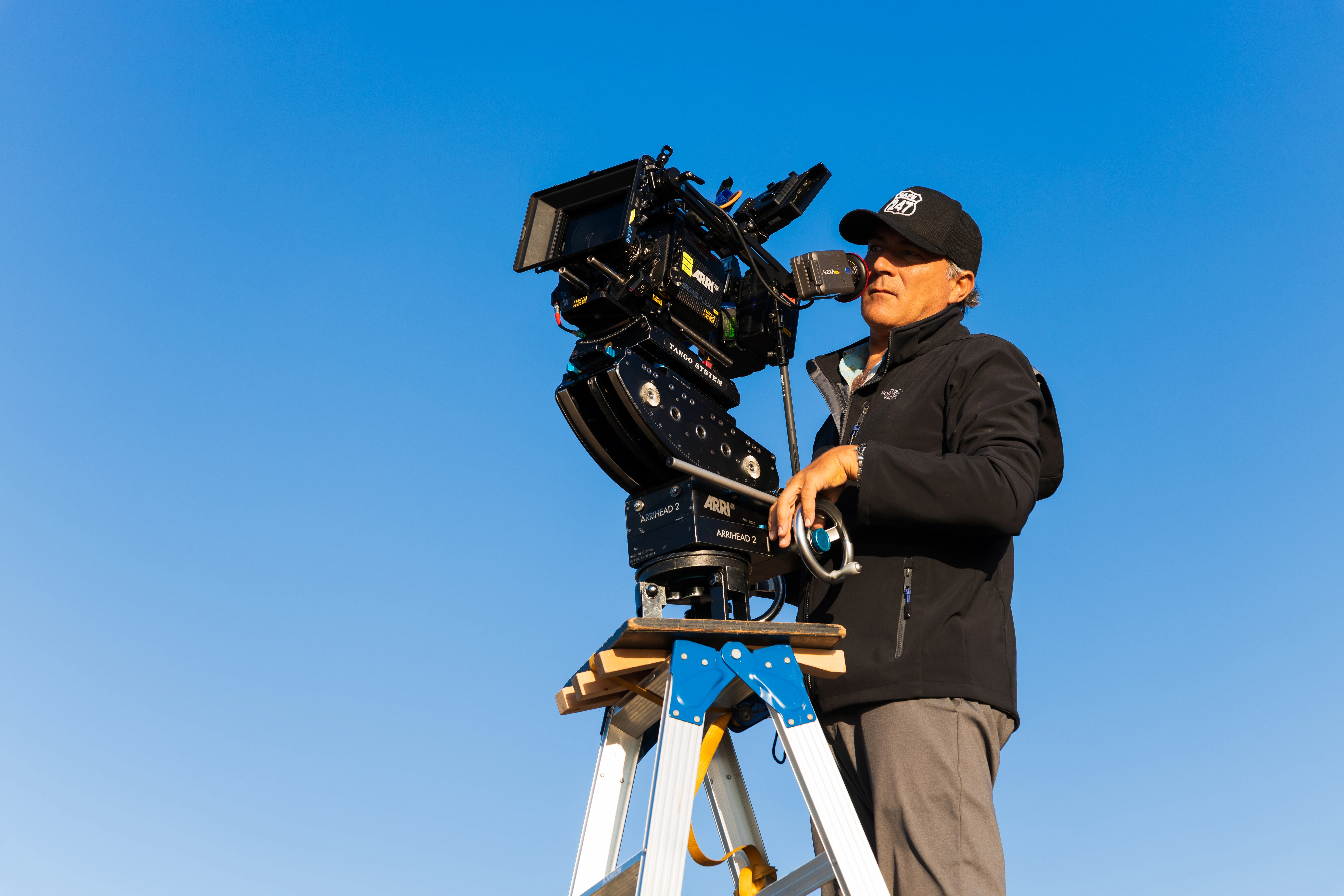 A camera operator filming outdoors beneath a clear blue sky.